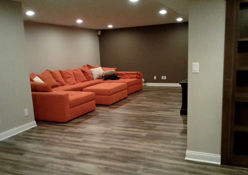 Living room with brown walls, wood flooring, and an orange sectional sofa. Ceiling lights illuminate the space.