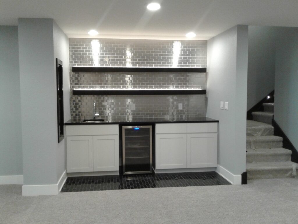 Modern basement kitchenette with white cabinets, black countertop, silver subway tile backsplash, shelves, and mini fridge.