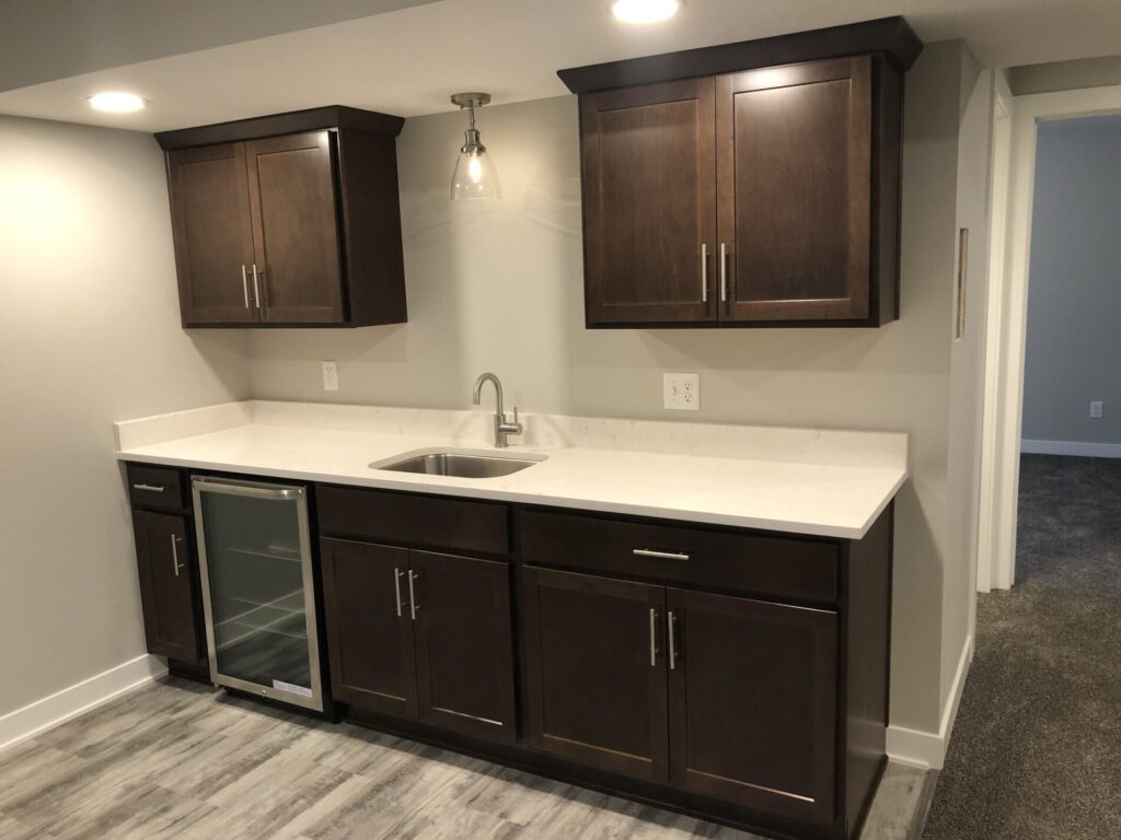 Basement wet bar with dark wood cabinets, a small sink, and a mini-fridge. The countertop is white, and flooring is vinyl.