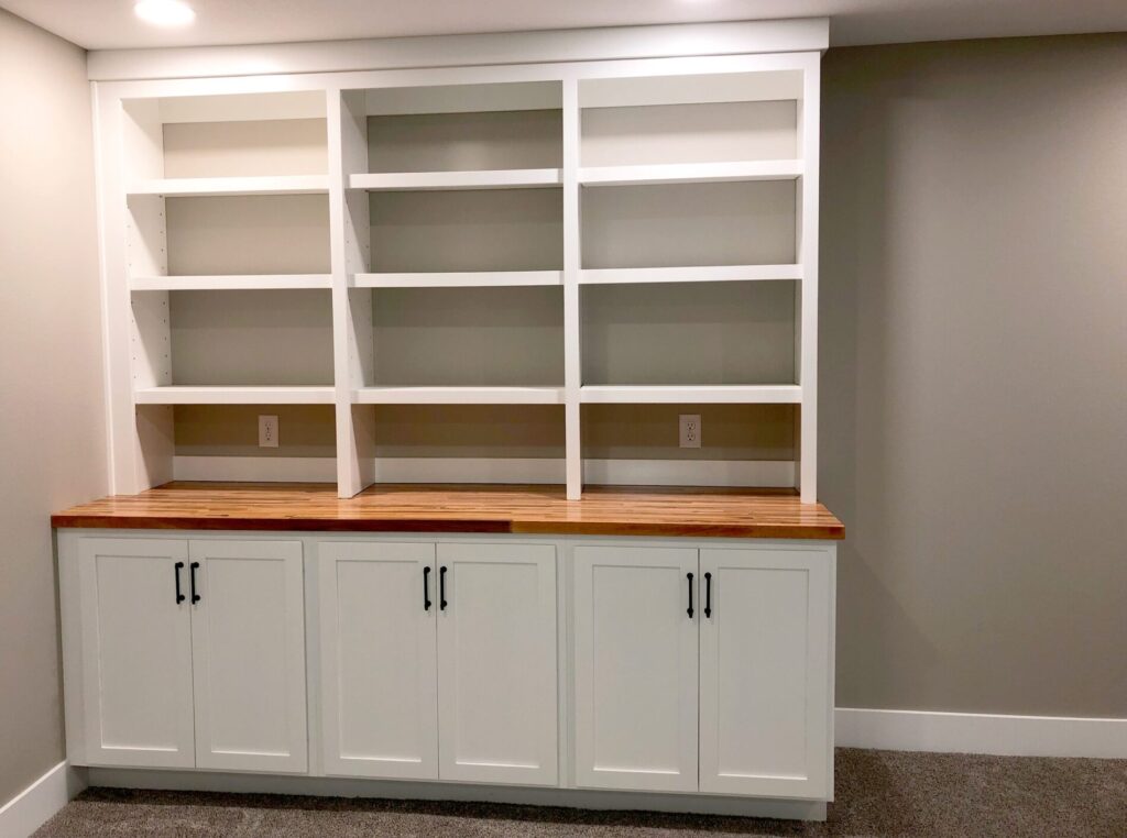 White built-in shelves with a wooden countertop and lower cabinet doors in a room with gray walls and carpeted floor.