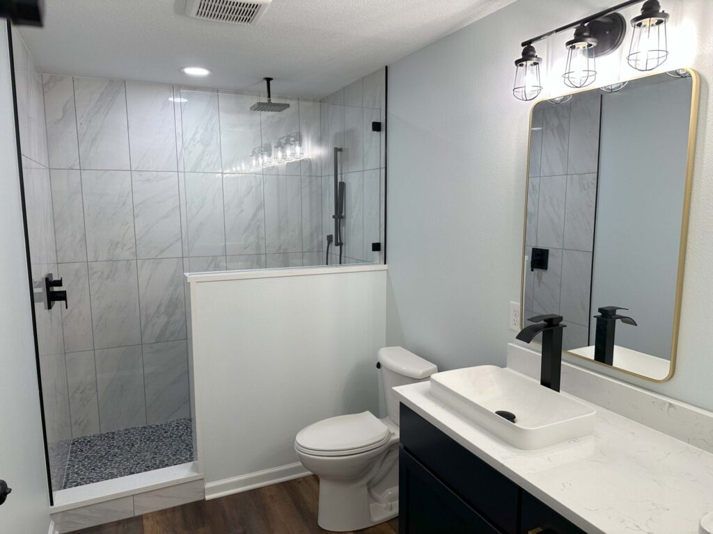 Modern bathroom with a glass-enclosed shower, white sink on a black vanity, toilet, and wood flooring.