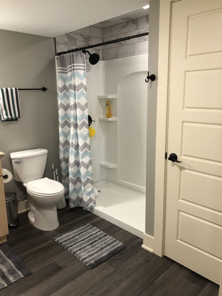 Bathroom with a shower, patterned curtain, toilet, and striped mats on a dark floor. Towel hangs on a black rod.