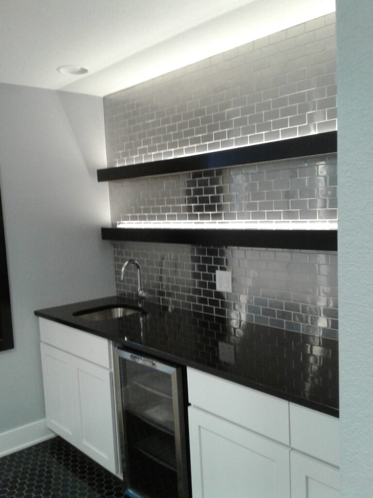 Modern kitchen area with white cabinets, black countertop, metal subway tile backsplash, and built-in lighting above shelves.