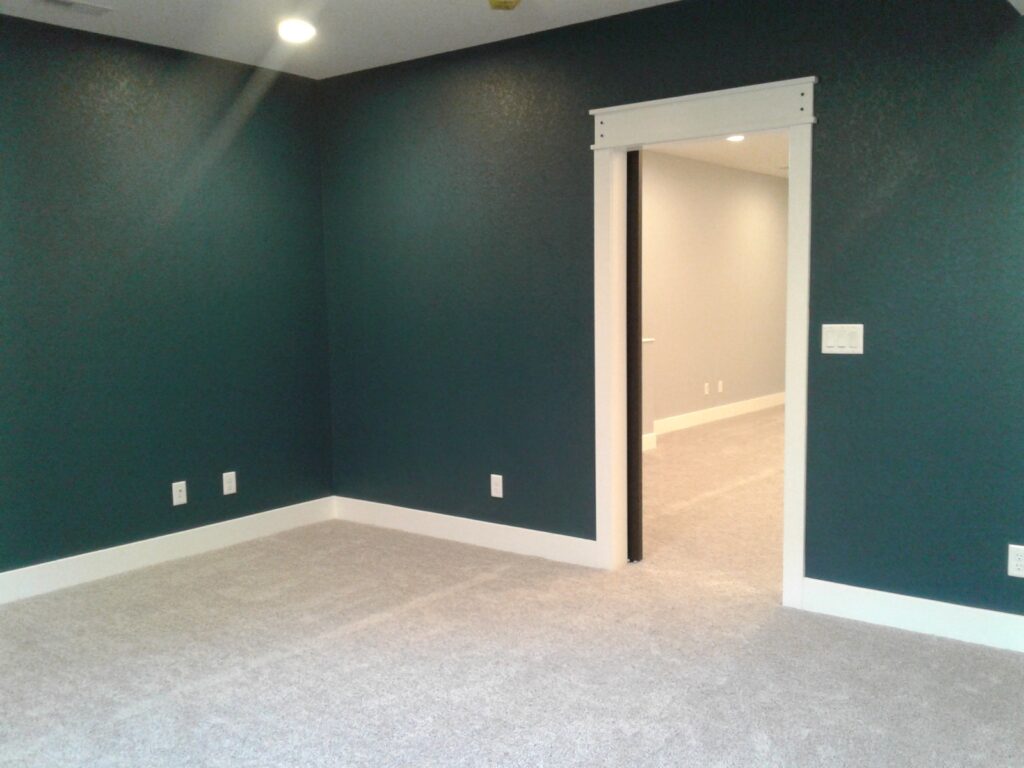 Empty room with teal walls and gray carpet, featuring a doorway leading to another room with a beige wall.