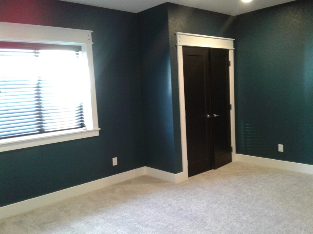 Empty room with teal walls, white trim, a window with blinds, and a black double-door closet on a carpeted floor.