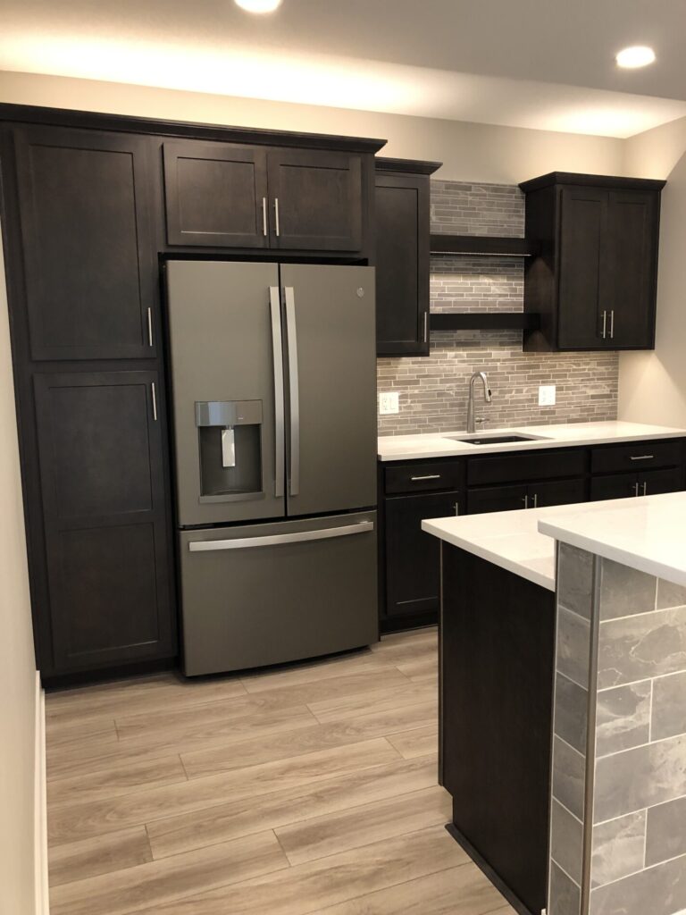 Modern kitchen with dark cabinets, stainless steel refrigerator, tile backsplash, and light wooden floor.