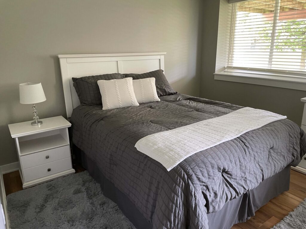 A bedroom with a neatly made bed featuring gray bedding, two white pillows, a nightstand with a lamp, and a window with blinds.