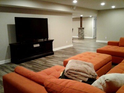 A basement living room with orange sofas, a large TV on a stand, wood flooring, and a column with stone decor.