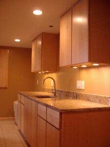 A modern kitchen with wooden cabinets, marble countertops, and a stainless steel faucet, illuminated by built-in ceiling lights.
