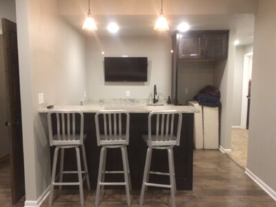 Basement bar area with three metal stools, a granite countertop, a wall-mounted TV, and overhead lights.