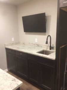 A small kitchenette with dark cabinets, a white countertop, a sink with a black faucet, and a wall-mounted TV.