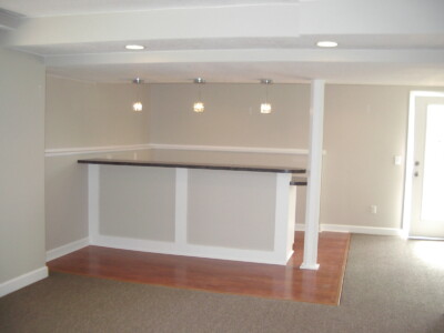 A basement room with a small bar, wooden countertop, pendant lights, gray walls, carpeted area, and a door to the right.