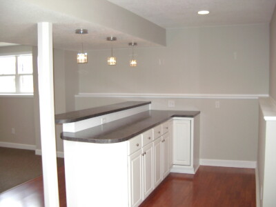 A basement kitchenette with white cabinets, a dark countertop, pendant lights, and wooden flooring.