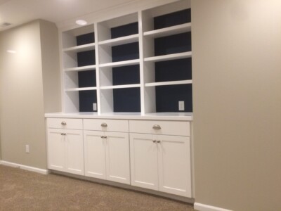 White built-in shelving unit with open shelves and closed cabinets against a beige wall, set on a carpeted floor.