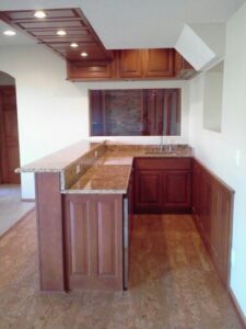 Modern home bar with wooden cabinets, granite countertops, sink, and recessed lighting under a coffered ceiling.
