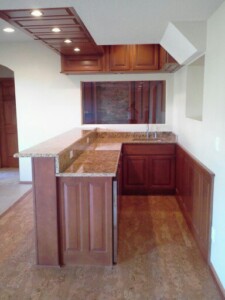 Modern home bar with wooden cabinets, granite countertops, sink, and recessed lighting under a coffered ceiling.