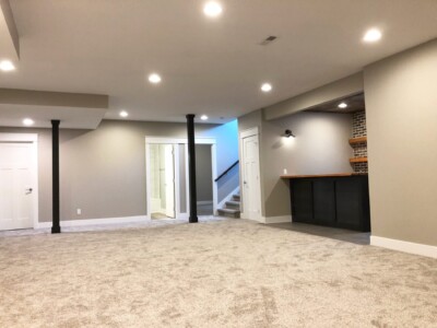 Finished basement with carpeted floor, recessed lighting, a small bar area with shelves, and a staircase in the background.