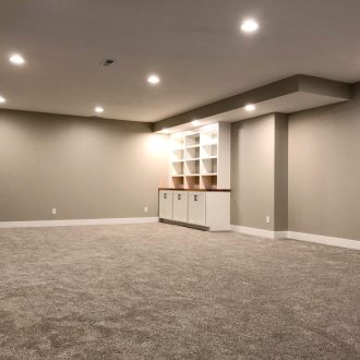 A spacious, carpeted basement with beige walls, recessed lighting, and built-in shelves and cabinets on the far wall.