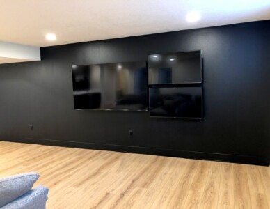 Black wall with three mounted flat-screen TVs of different sizes, set on a light wood floor with recessed lighting above.