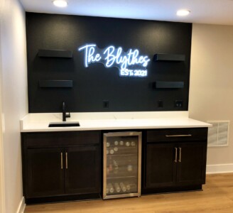 A home bar with dark cabinets, a mini fridge, and a neon sign reading "The Blythes EST. 2021" on a black wall.