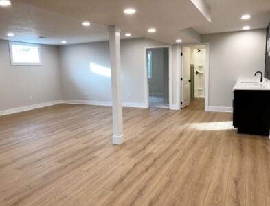 Spacious empty basement with light wood flooring, recessed lighting, a small kitchenette, and an open bathroom entrance.