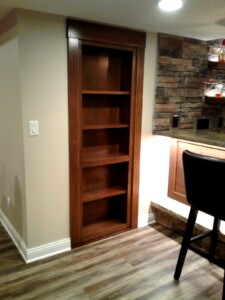 Wooden bookshelf integrated into a wall next to a bar area with stone accents and a bar stool on hardwood flooring.