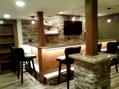 Home bar area with stone accents, a wooden counter, high chairs, wall-mounted TV, and overhead lights in a finished basement.