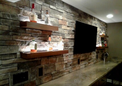 Stone wall with illuminated shelves displaying various liquor bottles, a large flat-screen TV, and a countertop with a sink.