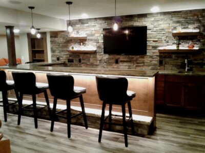 A home bar with four stools, a stone wall, shelves, a countertop, and a mounted TV, illuminated by hanging lights.