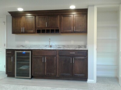 Wooden kitchen cabinets with a stone countertop, a built-in wine rack, a small fridge, and an adjacent empty pantry.