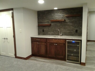 A modern kitchenette features dark wood cabinets, a mini-fridge, and stone backsplash with shelves, under recessed lighting.