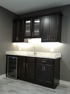 Dark wood kitchenette with upper and lower cabinets, marble countertop, small sink, and mini fridge on light wood flooring.