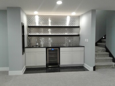 Modern basement kitchenette with white cabinets, black countertop, silver subway tile backsplash, shelves, and mini fridge.