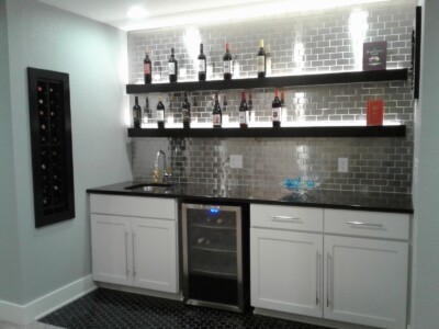 Home bar with white cabinets, a small fridge, and shelves displaying wine bottles against a tiled backsplash.