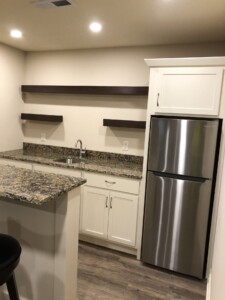 Compact kitchen with granite countertops, shelves, a sink, and stainless steel fridge. Cream cabinets and wood flooring.