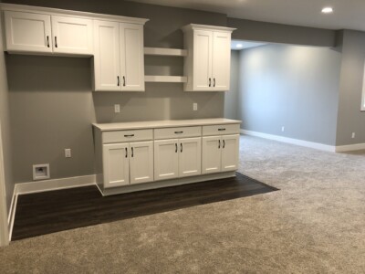 A basement room with white cabinets, beige carpet, and a gray wall. There is a small area with dark wood flooring.