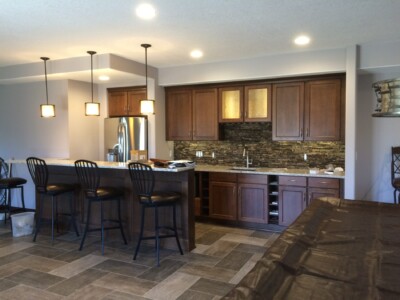 Modern kitchen with a stone backsplash, wooden cabinets, a stainless steel fridge, pendant lights, and four black bar stools.
