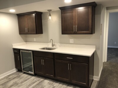 Basement wet bar with dark wood cabinets, a small sink, and a mini-fridge. The countertop is white, and flooring is vinyl.