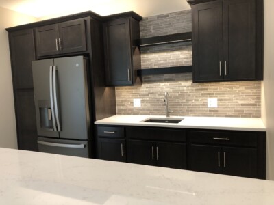 Modern kitchen with dark cabinets, stainless steel fridge, and light gray tile backsplash above a white countertop and sink.