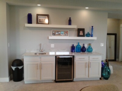 Room with a wet bar featuring white cabinets, a mini fridge, a sink, and shelves displaying blue glass bottles and photos.