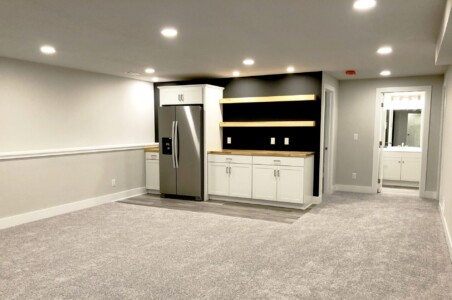 A modern, carpeted basement features a kitchenette with white cabinets, open shelves, and a stainless steel refrigerator.
