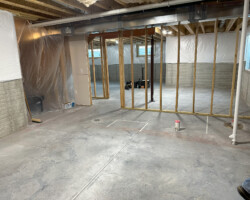 Partially finished basement with exposed wooden framing, concrete floor, and plastic sheeting on walls and ceiling.