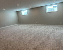 An empty basement room with gray walls, beige carpet, and two small rectangular windows near the ceiling.