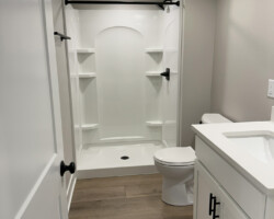 A modern bathroom with white shower, toilet, sink, and wood flooring. White walls and black fixtures complete the design.