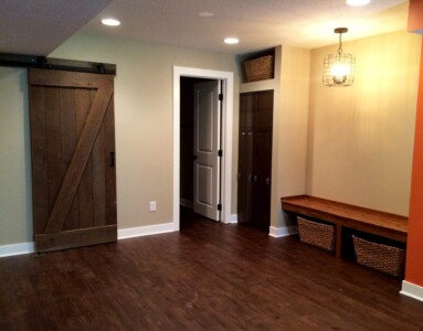 Basement room with wooden floor, barn door, lockers, bench with baskets, and light fixture. Door to another room visible.