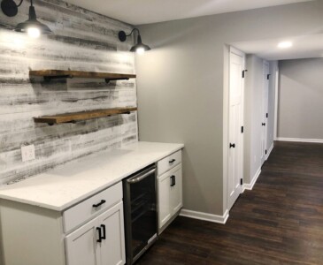Modern basement with wood floors, white cabinetry, mini-fridge, rustic shelves, and whitewashed accent wall with lighting.
