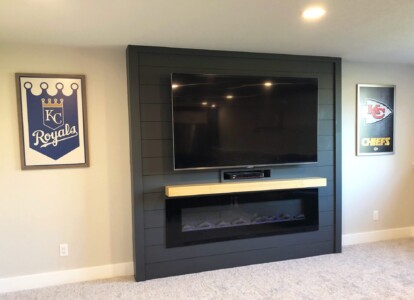 Wall-mounted TV with a wooden fireplace below, flanked by KC Royals and Chiefs framed posters.
