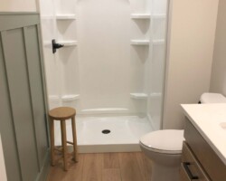 Bathroom with a walk-in shower, wooden stool, white toilet, and wooden vanity on light wood flooring.
