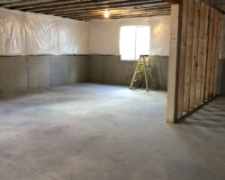 Unfinished basement with exposed beams, a ladder, and plastic-covered walls. Concrete floor and a single window visible.