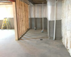 Unfinished basement with exposed wooden beams, pipes, insulation, and a yellow ladder against a concrete wall.